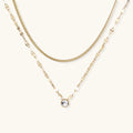 Layered gold necklace with herringbone and link chains, small bezel-set crystal pendant on cream background