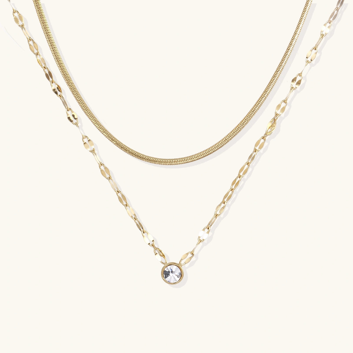 Layered gold necklace with herringbone and link chains, small bezel-set crystal pendant on cream background