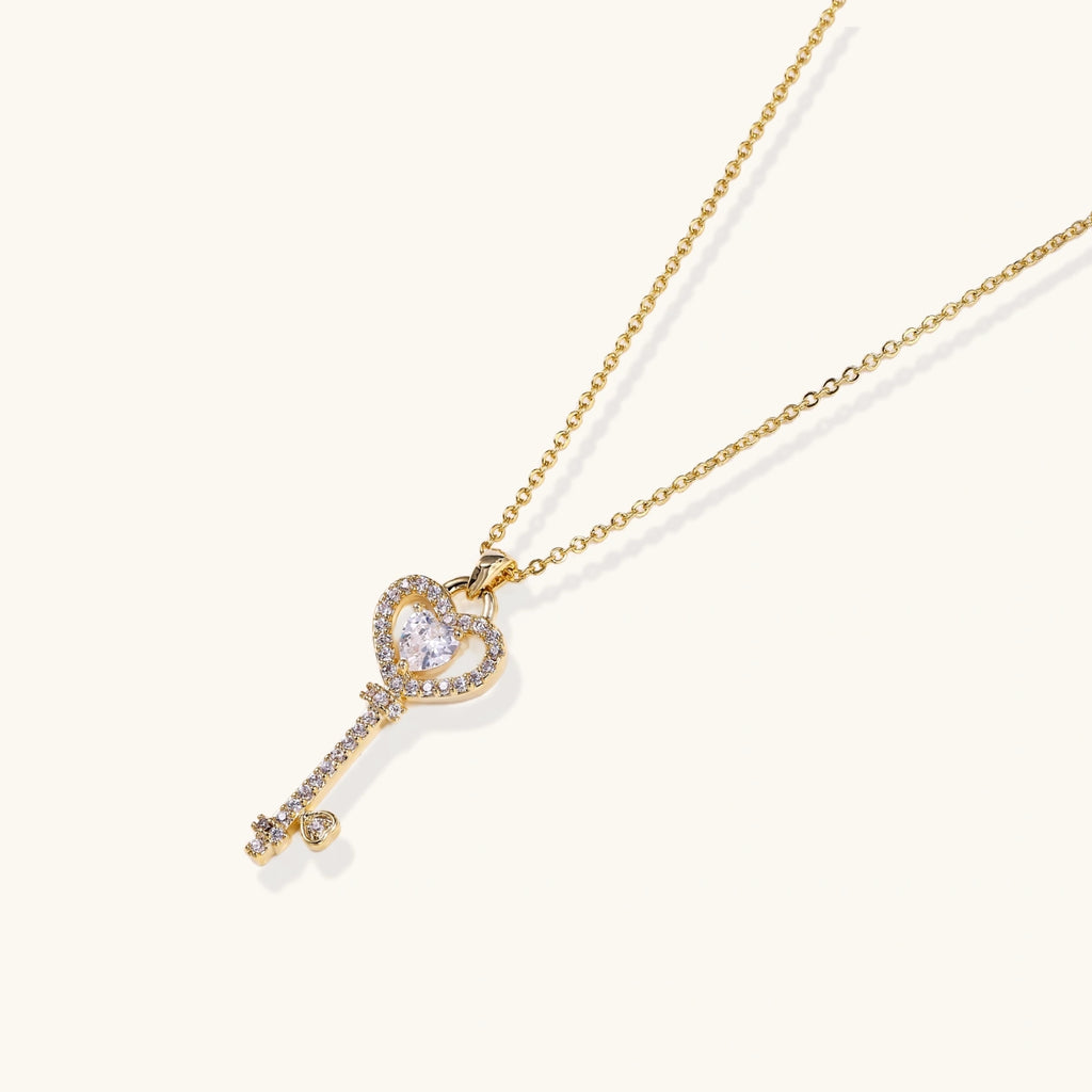 Delicate gold key pendant with heart-shaped bow and crystal pavé detailing on fine chain against cream background.