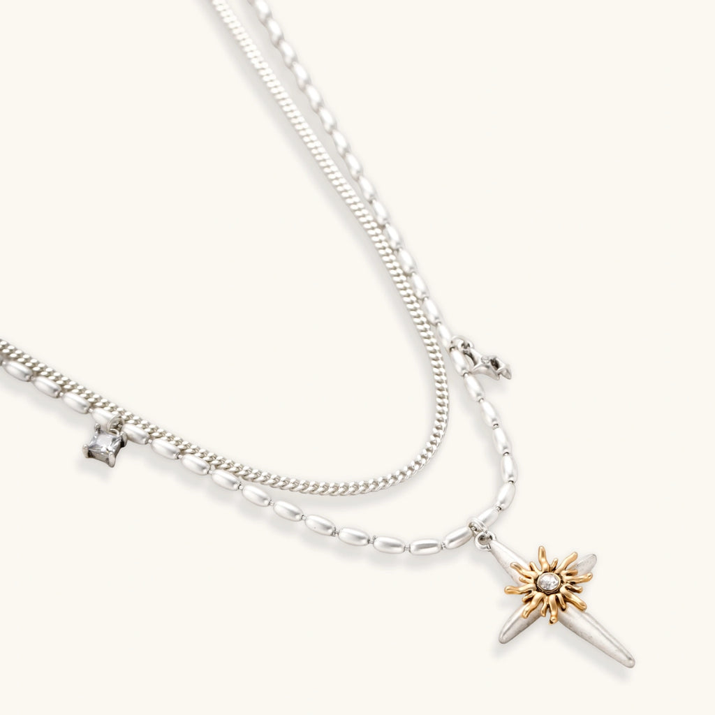 Layered silver-toned necklace set with cross pendant and gold sun charm detail, matching drop earrings on white background.