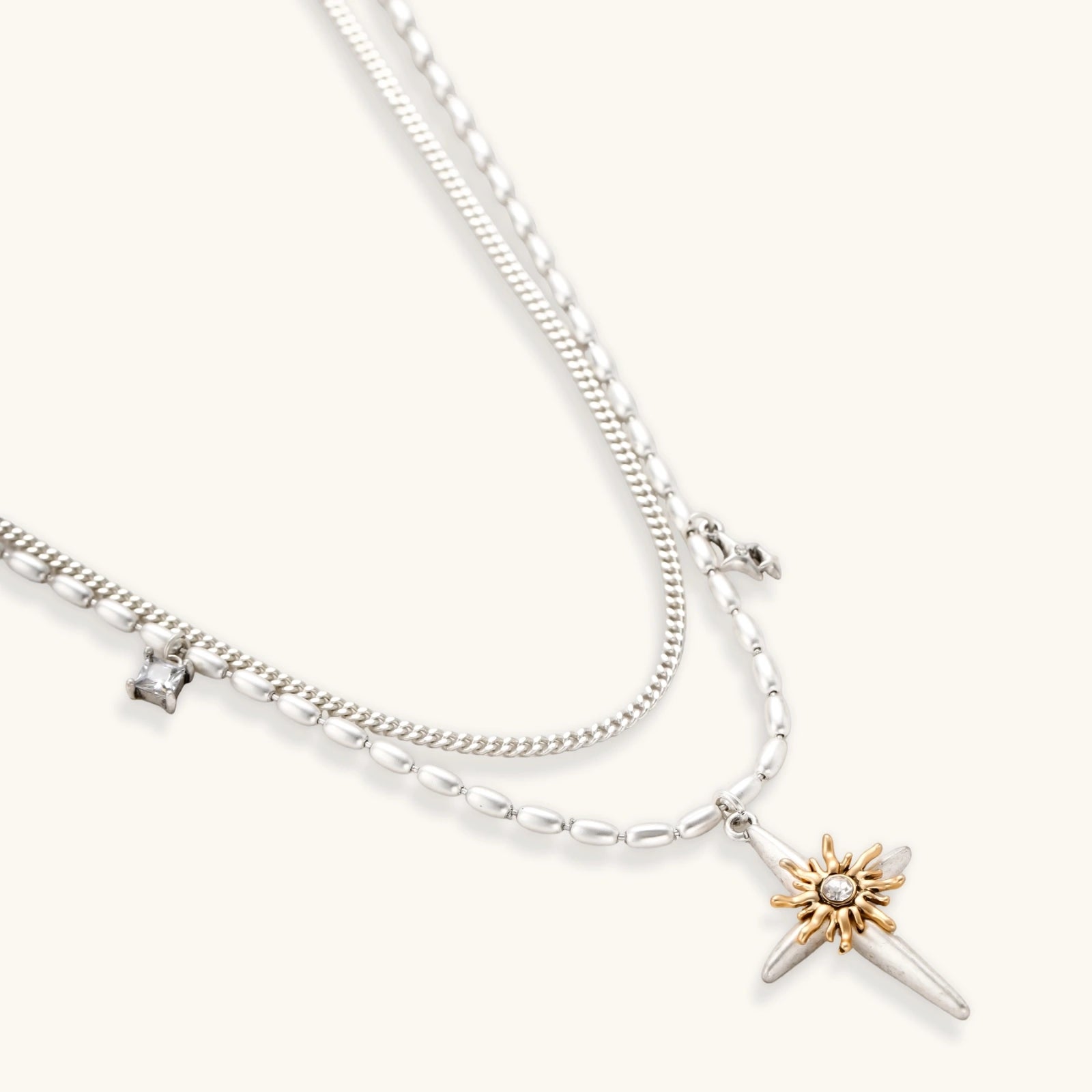 Layered silver-toned necklace set with cross pendant and gold sun charm detail, matching drop earrings on white background.