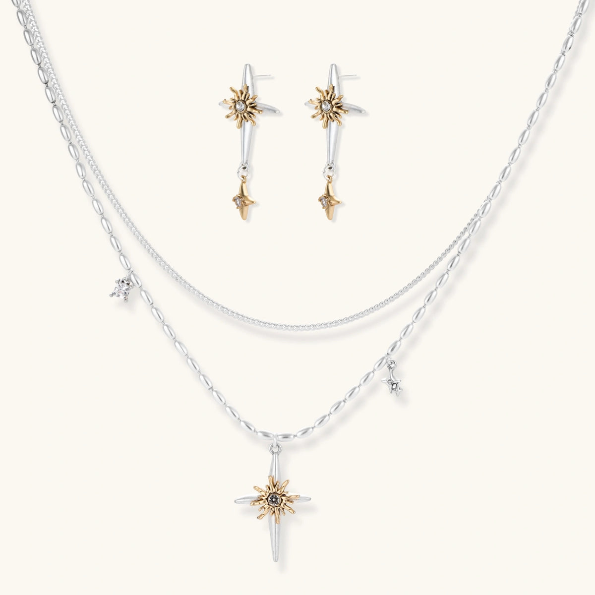 Layered silver-toned necklace set with cross pendant and gold sun charm detail, matching drop earrings on white background.