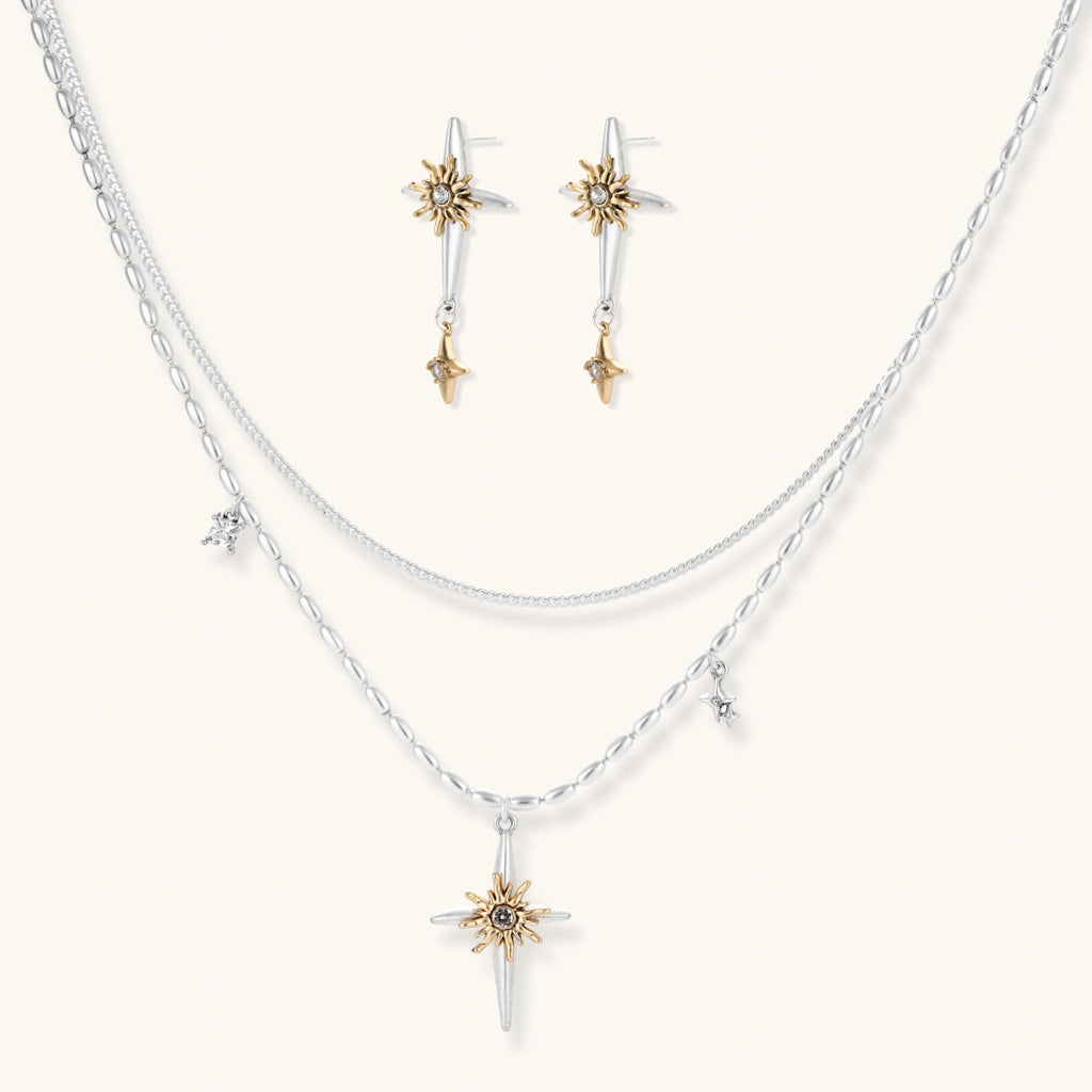 Layered silver-toned necklace set with cross pendant and gold sun charm detail, matching drop earrings on white background.