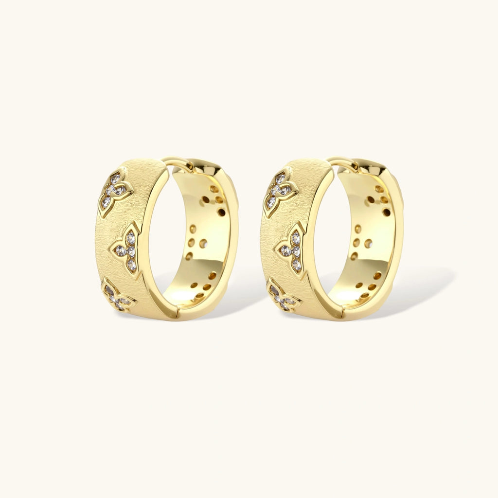 Gold-plated hoop earrings with textured surface and raised floral motifs set with clear zirconia stones on cream background.